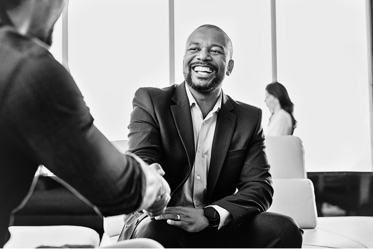 Business man smiling broadly while shaking hands with another man in a long sleeved shirt